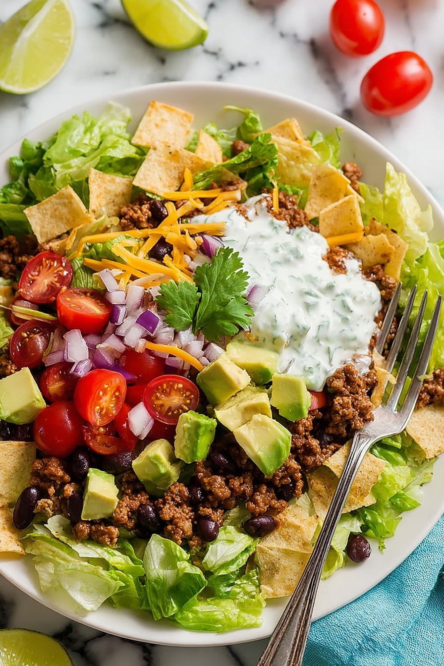 The dish shows a white plate filled with layers starting with fresh green lettuce leaves at the bottom. On top, there are scattered pieces of light beige tortilla chips mixed with cooked ground meat and black beans. Bright red halved cherry tomatoes and small cubes of green avocado are spread across the plate. There are thin strips of orange cheese and small chunks of purple onion scattered unevenly. Near the center, there is a dollop of white creamy sauce with green herbs, topped with a single cilantro leaf. A silver fork rests on the upper left edge of the plate, all placed on a white marbled surface with a few whole cherry tomatoes and lime wedges visible nearby. photo taken with an iphone --ar 2:3 --v 7