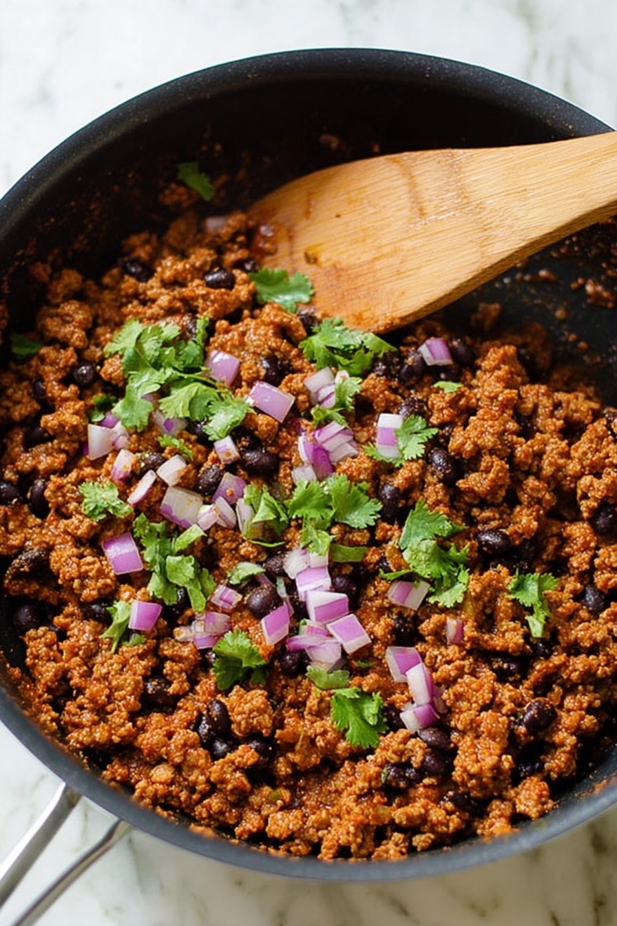 The image shows a black pan filled with cooked ground meat mixed with black beans. The meat mixture is a reddish-brown color with a crumbly texture, and it is topped with small pieces of purple and white chopped onion and fresh green cilantro leaves scattered on top. A wooden spatula is resting inside the pan on the left side. The pan is placed on a white marbled surface. photo taken with an iphone --ar 2:3 --v 7